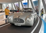 A classic silver sports car with a prominent Mercedes-Benz emblem on its grille is displayed on a curved platform inside a modern showroom. Several people are observing the car, including a man in a yellow sweater and blue jeans who is standing close by, wearing a face mask. The background features several large poster displays and other vintage cars.