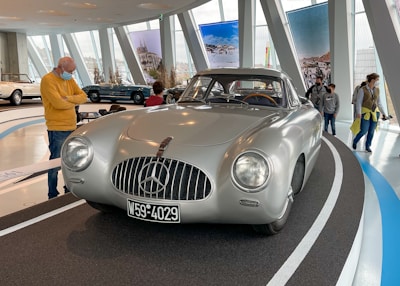 A classic silver sports car with a prominent Mercedes-Benz emblem on its grille is displayed on a curved platform inside a modern showroom. Several people are observing the car, including a man in a yellow sweater and blue jeans who is standing close by, wearing a face mask. The background features several large poster displays and other vintage cars.