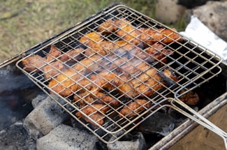 A barbecue grill is shown with pieces of marinated chicken roasting over hot charcoal. The meat appears to be well-cooked with a crispy, browned texture. Smoke is wafting up from the coals, adding to the rustic outdoor cooking scene.