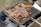 A barbecue grill is shown with pieces of marinated chicken roasting over hot charcoal. The meat appears to be well-cooked with a crispy, browned texture. Smoke is wafting up from the coals, adding to the rustic outdoor cooking scene.