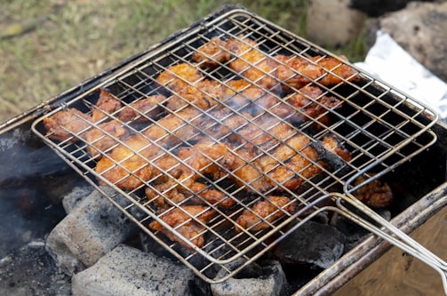 A barbecue grill is shown with pieces of marinated chicken roasting over hot charcoal. The meat appears to be well-cooked with a crispy, browned texture. Smoke is wafting up from the coals, adding to the rustic outdoor cooking scene.