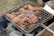 A barbecue grill is shown with pieces of marinated chicken roasting over hot charcoal. The meat appears to be well-cooked with a crispy, browned texture. Smoke is wafting up from the coals, adding to the rustic outdoor cooking scene.
