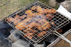 A barbecue grill is shown with pieces of marinated chicken roasting over hot charcoal. The meat appears to be well-cooked with a crispy, browned texture. Smoke is wafting up from the coals, adding to the rustic outdoor cooking scene.