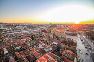 An aerial view of a cityscape featuring red-tiled rooftops, historical buildings, and a river running through the city. The sun is setting in the background, casting a warm glow over the scene. A main road is visible with several cars and pedestrians.