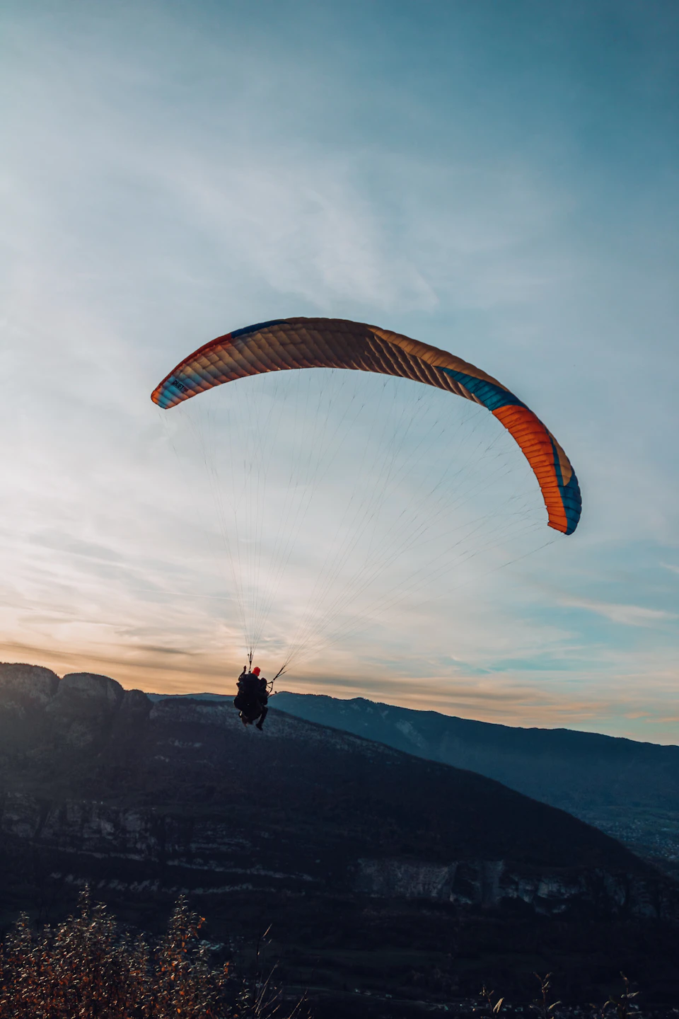 The image depicts a person paragliding over a mountain range at sunset. The paraglider is suspended in mid-air, with their body angled towards the right side of the frame and their feet slightly above the ground. The parachute they are attached to has an orange and blue color scheme, contrasting against the warm hues of the setting sun.

The backdrop features a breathtaking view of mountains under a clear sky, with the sun casting long shadows and highlighting the contours of the landscape.