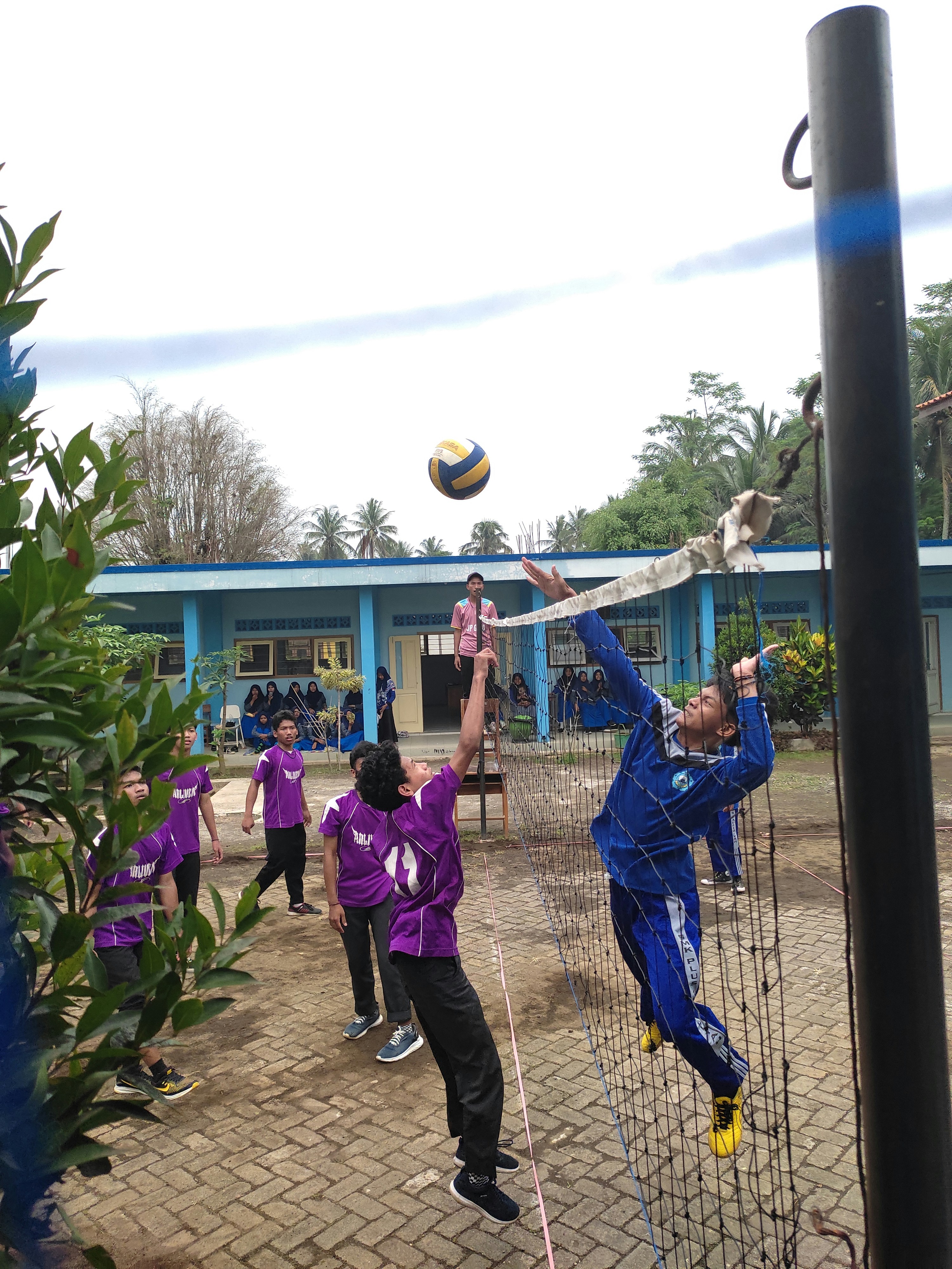 A group of young people are playing volleyball on a court near a blue building. One player, dressed in blue, is jumping to hit the ball over the net while other players in purple uniforms watch and prepare to engage. The court is outdoors, surrounded by greenery, with a clear sky above.