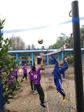 A group of young people are playing volleyball on a court near a blue building. One player, dressed in blue, is jumping to hit the ball over the net while other players in purple uniforms watch and prepare to engage. The court is outdoors, surrounded by greenery, with a clear sky above.