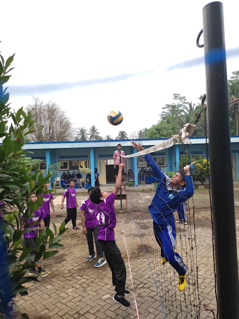 A group of young people are playing volleyball on a court near a blue building. One player, dressed in blue, is jumping to hit the ball over the net while other players in purple uniforms watch and prepare to engage. The court is outdoors, surrounded by greenery, with a clear sky above.