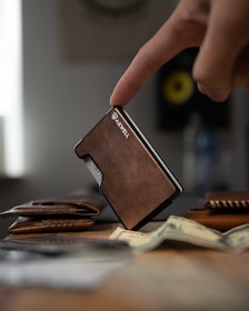 A finger gently rests on a sleek, brown leather wallet standing upright on a table. In the foreground, a stack of cash is slightly out of focus, adding depth to the scene. Other leather accessories are spread out, contributing to a rustic and organized setup. In the background, a blurred circular object with a yellow center is visible, suggesting a modern, tech-savvy environment.