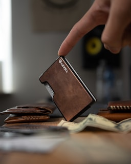 A finger gently rests on a sleek, brown leather wallet standing upright on a table. In the foreground, a stack of cash is slightly out of focus, adding depth to the scene. Other leather accessories are spread out, contributing to a rustic and organized setup. In the background, a blurred circular object with a yellow center is visible, suggesting a modern, tech-savvy environment.