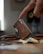 A finger gently rests on a sleek, brown leather wallet standing upright on a table. In the foreground, a stack of cash is slightly out of focus, adding depth to the scene. Other leather accessories are spread out, contributing to a rustic and organized setup. In the background, a blurred circular object with a yellow center is visible, suggesting a modern, tech-savvy environment.
