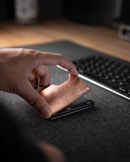 A hand is holding a slim, copper-colored metallic object with the brand name 'AXWELL' engraved on it, positioned above a desk mat. In the background, a portion of a computer keyboard is visible along the edge of a wooden table.