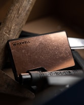 A close-up view of a copper-colored metallic card with the brand logo 'Axwell' displayed on its surface. The card is situated among tools, including a handle with 'U.N.T-D' engraved on it and a wooden plank. Dim lighting accentuates the textures and provides a warm, rustic ambiance.