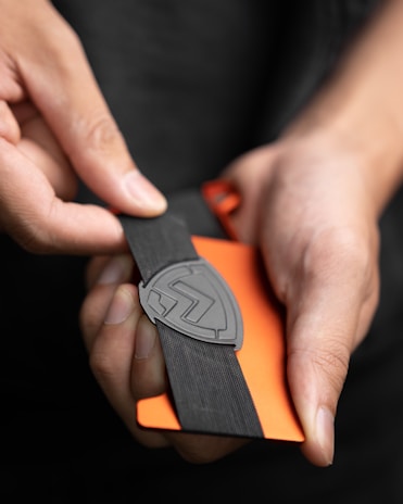 Hands are holding a black elastic band with a logo on it, wrapped around an orange card or device. The image focuses on the detailed texture of the elastic band and the grip of the fingers.