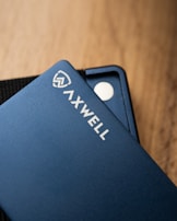 A close-up shot of a sleek blue wallet with the branding 'AXWELL' on its surface. The wallet rests on a wooden surface, with part of its interior design visible, including a small metallic disc.