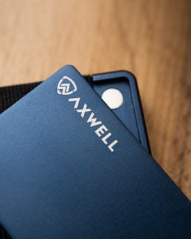 A close-up shot of a sleek blue wallet with the branding 'AXWELL' on its surface. The wallet rests on a wooden surface, with part of its interior design visible, including a small metallic disc.