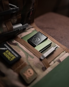 A military-style bag with a camouflage pattern contains a neatly arranged pocket organizer. Inside, there's a green metallic item with a black clip featuring an emblem. The bag has patches, including one that reads 'KNIFE ADDICT'. Various metal tools are tucked into the organizer, adding to the tactical aesthetic.