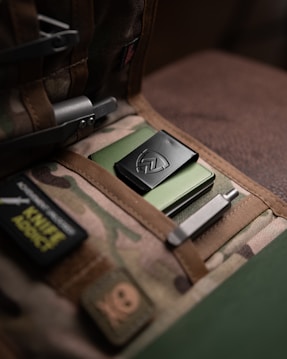 A military-style bag with a camouflage pattern contains a neatly arranged pocket organizer. Inside, there's a green metallic item with a black clip featuring an emblem. The bag has patches, including one that reads 'KNIFE ADDICT'. Various metal tools are tucked into the organizer, adding to the tactical aesthetic.