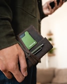 A close-up of a person's hand holding a brown leather wallet with a green card partially visible inside. A pen is tucked into the strap of the wallet. The person is wearing a dark green jacket and appears to be standing indoors with a blurred background.