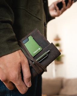 A close-up of a person's hand holding a brown leather wallet with a green card partially visible inside. A pen is tucked into the strap of the wallet. The person is wearing a dark green jacket and appears to be standing indoors with a blurred background.