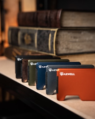 A lineup of sleek, minimalist wallets arranged on a surface in various colors, made by Axwell. These wallets are displayed in front of large, antiquated books with textured covers, creating a contrast between modern and traditional elements.