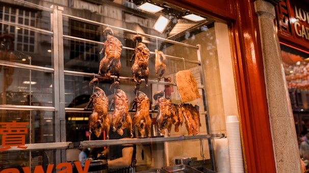 Several roasted ducks and a large piece of meat are displayed hanging inside a food stall with a glass window. The stall features red and steel tones, with reflections on the glass from the street outside.