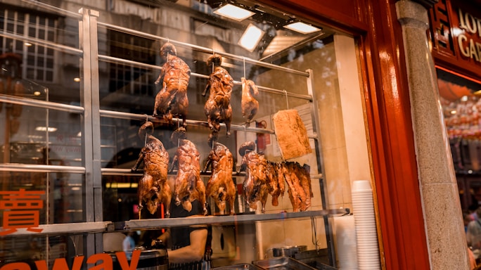 Several roasted ducks and a large piece of meat are displayed hanging inside a food stall with a glass window. The stall features red and steel tones, with reflections on the glass from the street outside.