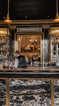 A bartender in a formal vest is working behind a bar stocked with various bottles of liquor. The bar has a luxurious design with a marble countertop and gold accents. Pendant lights hang from the ceiling, providing a warm ambiance. In the background, there is a large ornate mirror reflecting the elegant dining area with chandeliers and people seated at tables.