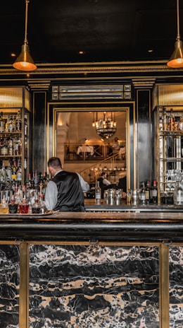 A bartender in a formal vest is working behind a bar stocked with various bottles of liquor. The bar has a luxurious design with a marble countertop and gold accents. Pendant lights hang from the ceiling, providing a warm ambiance. In the background, there is a large ornate mirror reflecting the elegant dining area with chandeliers and people seated at tables.