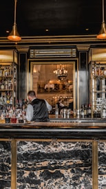 A bartender in a formal vest is working behind a bar stocked with various bottles of liquor. The bar has a luxurious design with a marble countertop and gold accents. Pendant lights hang from the ceiling, providing a warm ambiance. In the background, there is a large ornate mirror reflecting the elegant dining area with chandeliers and people seated at tables.