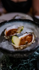 Two oysters served on a bed of ice in a decorative bowl. The oysters contain a red sauce and a slice of cucumber.