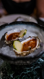 Two oysters served on a bed of ice in a decorative bowl. The oysters contain a red sauce and a slice of cucumber.