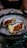 Two oysters served on a bed of ice in a decorative bowl. The oysters contain a red sauce and a slice of cucumber.