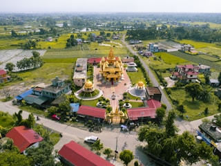 Aerial view of a golden temple with domes, surrounded by greenery and fields. The temple is situated in a village with roads and scattered residential houses. The landscape features lush green fields, trees, and distant hills under a clear sky.