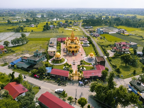 Aerial view of a golden temple with domes, surrounded by greenery and fields. The temple is situated in a village with roads and scattered residential houses. The landscape features lush green fields, trees, and distant hills under a clear sky.