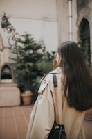 A woman in a flowing beige trench coat walking past a minimalist café with potted plants.