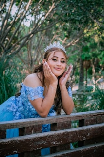 A young woman wearing an elegant blue gown adorned with floral details and a sparkling tiara poses gracefully, resting her face on her hands while leaning on a wooden railing in a lush, green park.