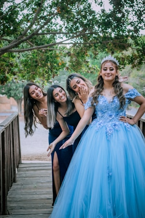 Four women are standing on a wooden bridge amidst a lush green background. One is wearing an elaborate blue gown with floral embellishments and a tiara, suggesting a celebratory occasion. The others are dressed in black outfits, smiling and leaning towards the woman in the gown.