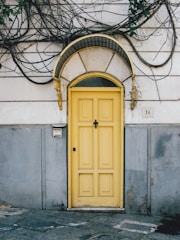 A vibrant yellow door with a decorative black and gold arch sits in the center of a worn, weathered wall. The wall is partially covered with creeping vines and a small number plaque displaying the number 14. The lower portion of the wall is painted gray, contrasting with the more faded, lighter upper area. A doorbell or intercom is mounted on the wall to the left of the door.
