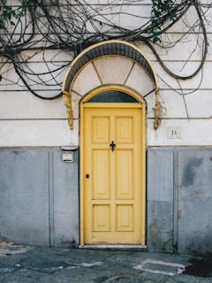 A vibrant yellow door with a decorative black and gold arch sits in the center of a worn, weathered wall. The wall is partially covered with creeping vines and a small number plaque displaying the number 14. The lower portion of the wall is painted gray, contrasting with the more faded, lighter upper area. A doorbell or intercom is mounted on the wall to the left of the door.