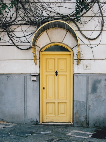 A vibrant yellow door with a decorative black and gold arch sits in the center of a worn, weathered wall. The wall is partially covered with creeping vines and a small number plaque displaying the number 14. The lower portion of the wall is painted gray, contrasting with the more faded, lighter upper area. A doorbell or intercom is mounted on the wall to the left of the door.