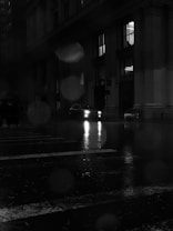 A nighttime scene featuring a dark, wet city street with reflections on the pavement. A car with illuminated headlights is partially visible in front of a large building with columns. The atmosphere is moody, with raindrops and a blurry effect present in the image.
