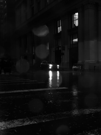 A nighttime scene featuring a dark, wet city street with reflections on the pavement. A car with illuminated headlights is partially visible in front of a large building with columns. The atmosphere is moody, with raindrops and a blurry effect present in the image.