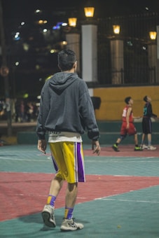 A person wearing a gray hoodie and yellow shorts is walking on an outdoor basketball court at night. The court is surrounded by lit street lamps, and a basketball game with other players in red and black jerseys is happening in the background.