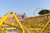 A large historic fort made of red sandstone is visible in the background, featuring prominent domes and an Indian flag atop. In the foreground, yellow security barricades are prominently placed, partially obscuring the view. To the right, a Ferris wheel with colorful cabins is visible next to a tree under a clear blue sky, while several people are seen standing near the barricades.