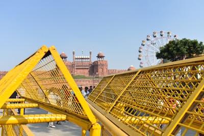 A large historic fort made of red sandstone is visible in the background, featuring prominent domes and an Indian flag atop. In the foreground, yellow security barricades are prominently placed, partially obscuring the view. To the right, a Ferris wheel with colorful cabins is visible next to a tree under a clear blue sky, while several people are seen standing near the barricades.