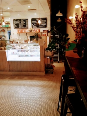 A cozy caf&eacute; setting with a wooden counter and stools on the right. A well-lit display case filled with pastries and confectioneries sits at the center. The walls have a white backsplash with menu boards hanging above. There are plants and flowers adding a touch of greenery.