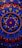 An intricate circular mandala pattern with vibrant colors including red, blue, gold, and pink. The design features concentric circles, floral motifs, and dots that create a symmetrical and ornate look. Elements radiate from the center and are repeated rhythmically, forming a harmonious and elaborate design.