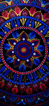 An intricate circular mandala pattern with vibrant colors including red, blue, gold, and pink. The design features concentric circles, floral motifs, and dots that create a symmetrical and ornate look. Elements radiate from the center and are repeated rhythmically, forming a harmonious and elaborate design.