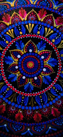 An intricate circular mandala pattern with vibrant colors including red, blue, gold, and pink. The design features concentric circles, floral motifs, and dots that create a symmetrical and ornate look. Elements radiate from the center and are repeated rhythmically, forming a harmonious and elaborate design.